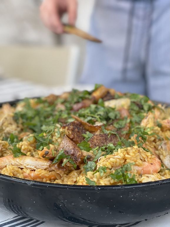 Paella in a large black pan, topped with herbs, with a person holding a wooden spoon.