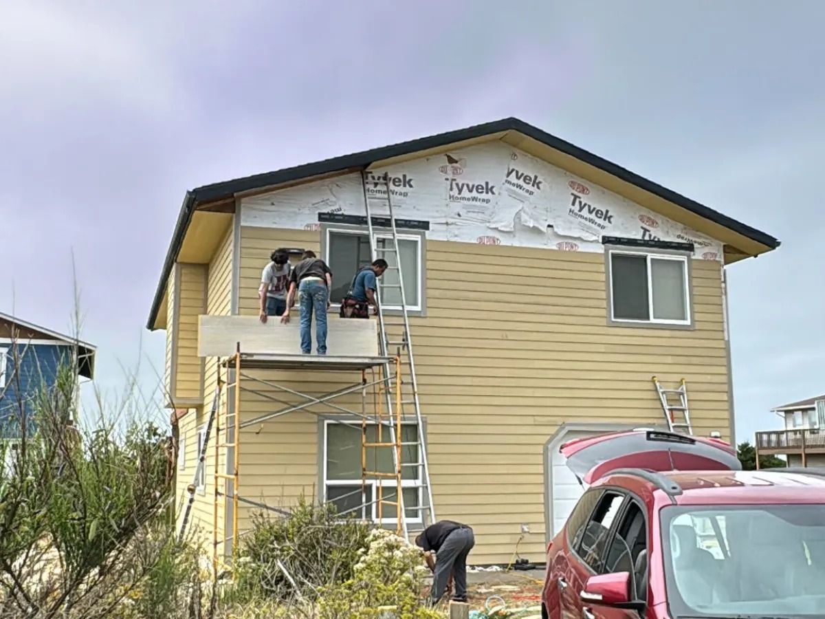 Workers installing siding on a two-story yellow house, using a scaffold. A car is parked nearby.