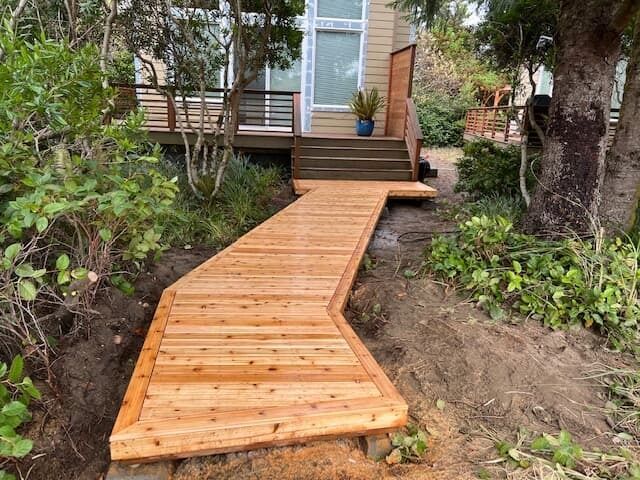 Wooden walkway leading to a house with steps and deck, surrounded by greenery.