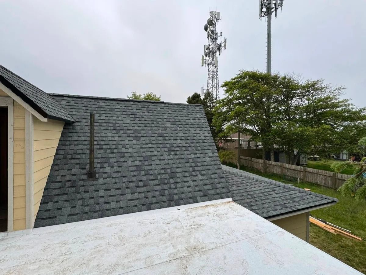 View of a dark gray shingled roof with a chimney and part of a cream-colored building.