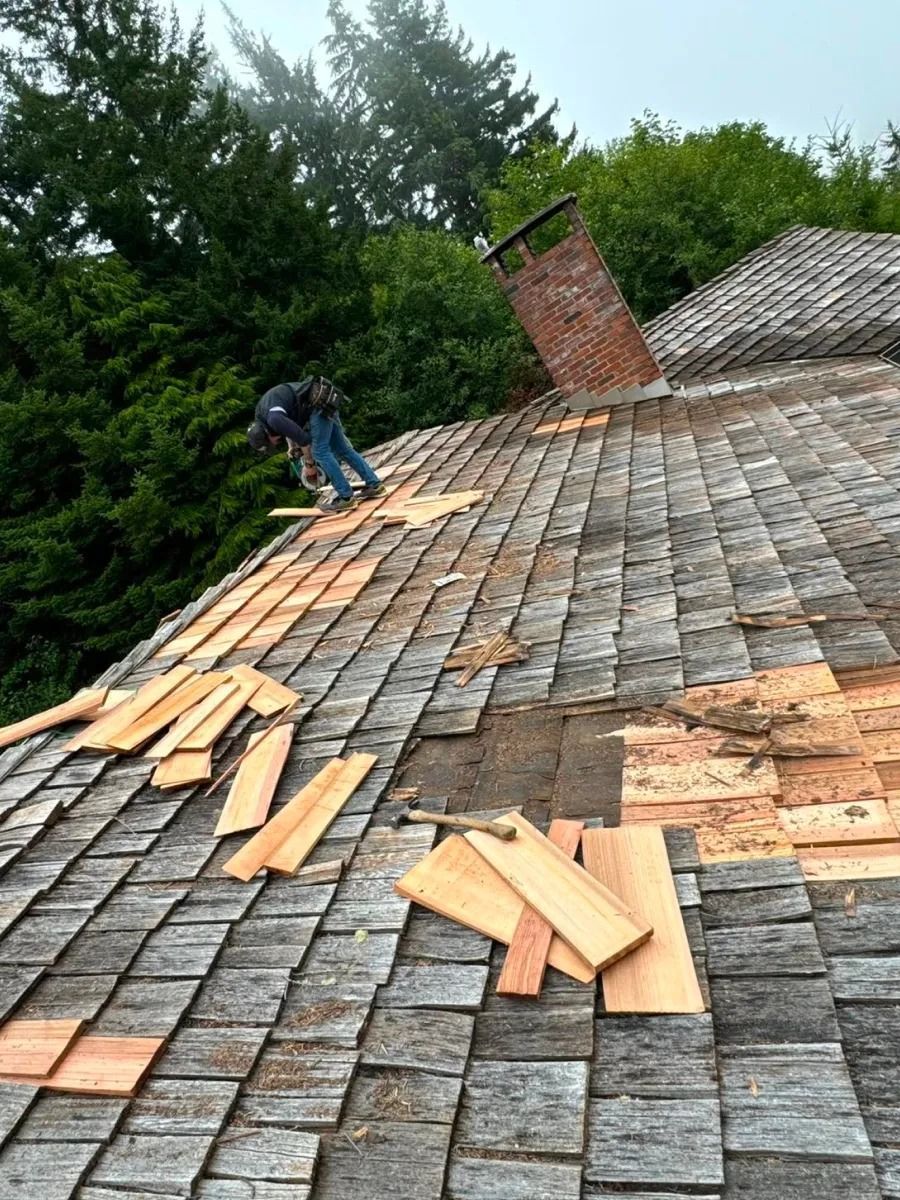 Workers replacing roof shingles on a house with a chimney; green trees in the background.