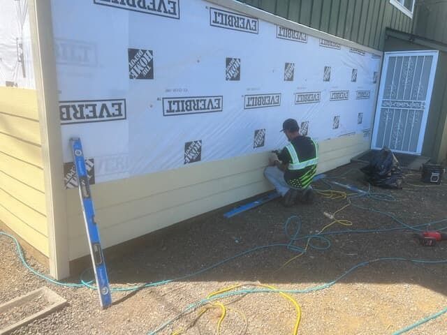 Man installing light yellow siding on a house exterior. White weather barrier visible.
