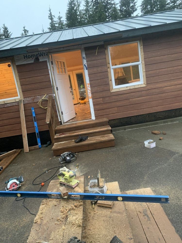 Construction site with brown siding and a metal roof. A doorway and window frame are visible, with tools in the foreground.