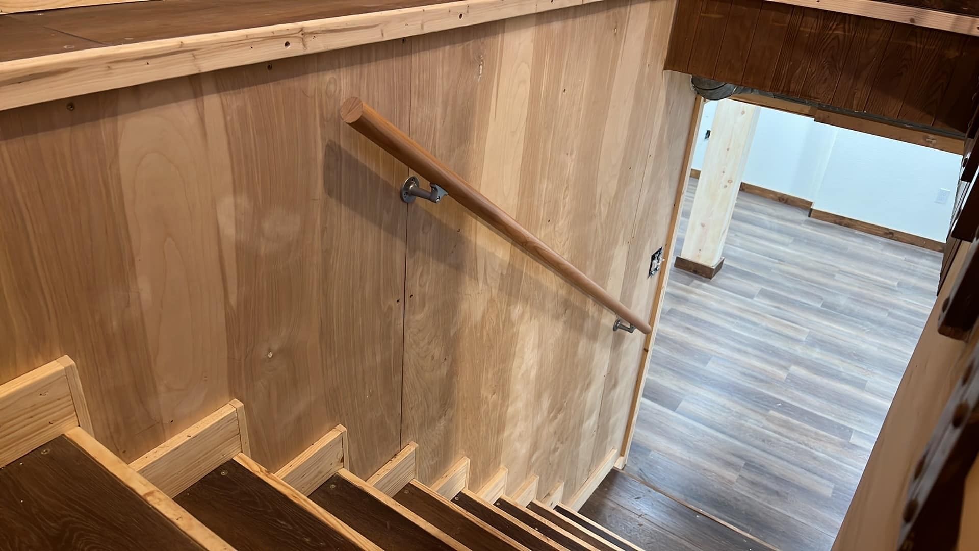 Wooden stairs with a handrail, leading downwards. The wall is light wood, and flooring is visible.