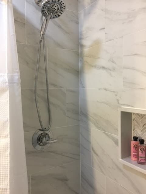 Shower with marble-patterned tile and a built-in shelf holding toiletries. Silver shower head and handheld.