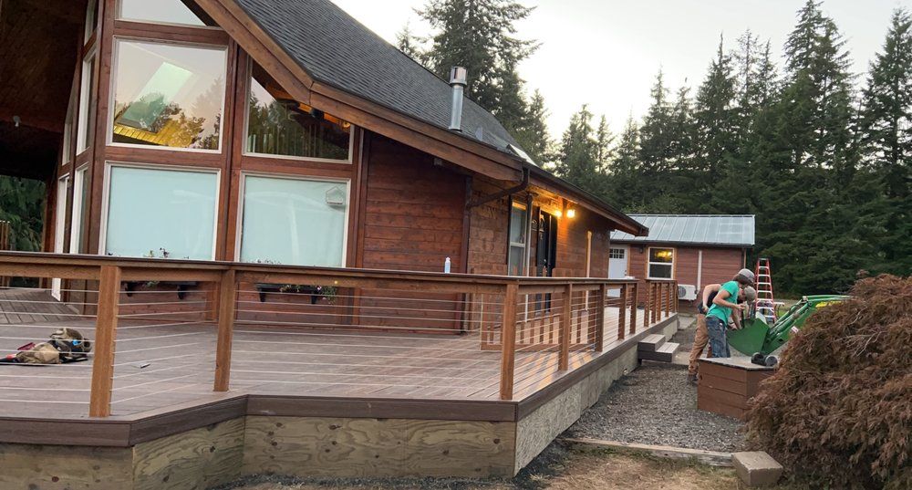 Wooden cabin with deck; people working, building in yard. Forest backdrop.