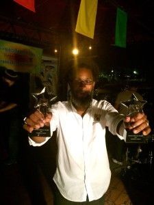 Man holding two star-shaped awards, smiling. Wearing a white shirt, in a dimly lit setting with colorful flags.