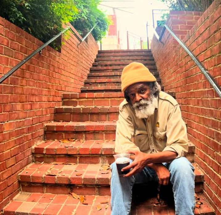 Mighty Joshua sitting on brick steps, holding a cup. He wears a yellow cap, light shirt, and jeans. Steps lead uphill.