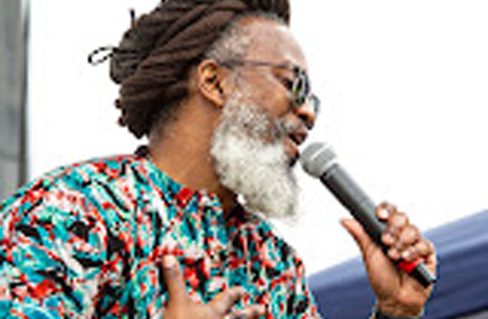 Mighty Joshua with dreadlocks and gray beard speaks into microphone, wearing patterned shirt and sunglasses.