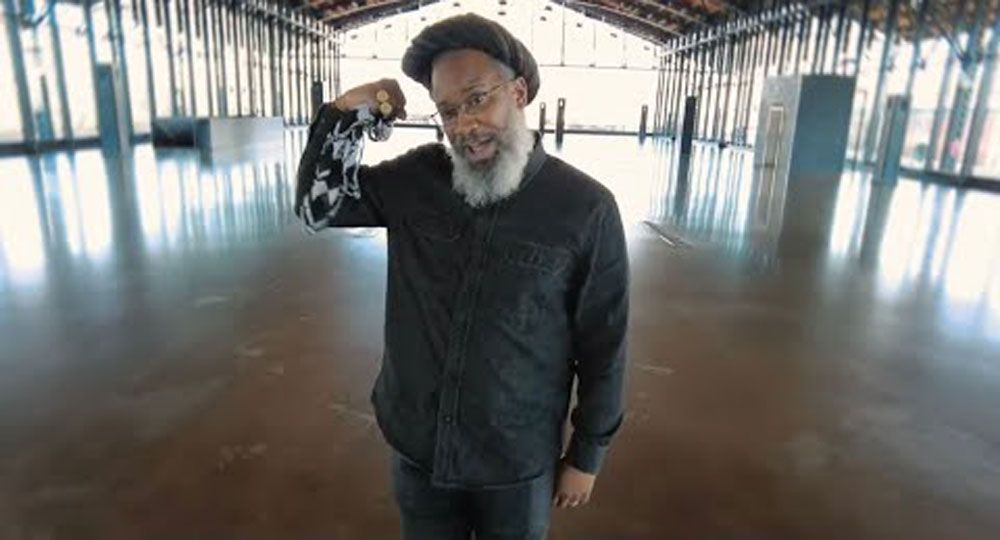 Mighty Joshua in black shirt and cap flexing arm, inside a large, empty, open-air building.