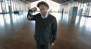 Mighty Joshua in black shirt and cap flexing arm, inside a large, empty, open-air building.