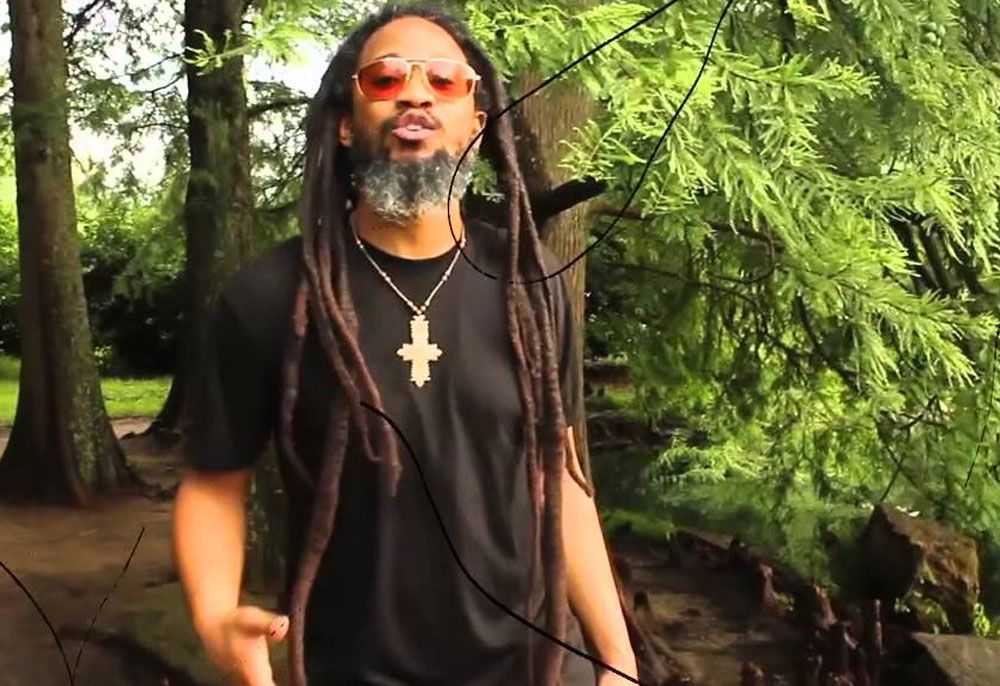 Mighty Joshua with dreadlocks and sunglasses in front of trees, wearing a black shirt and a cross necklace.