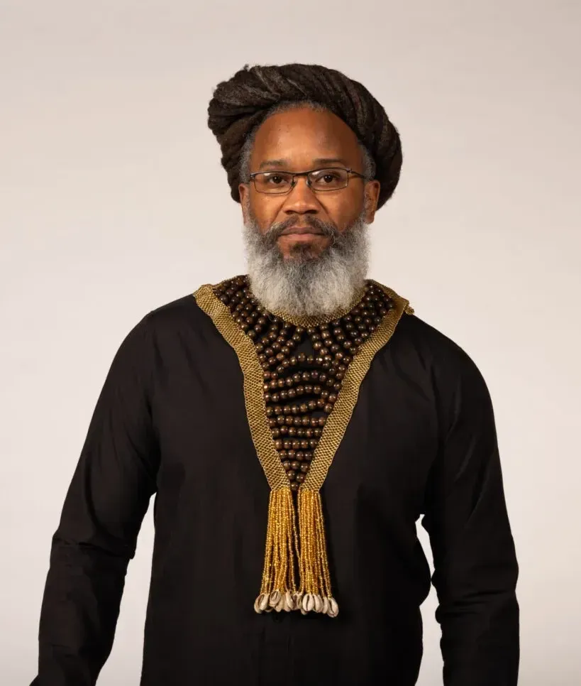 Mighty Joshua with dreadlocks, glasses, and a gray beard wearing a black shirt with ornate collar.