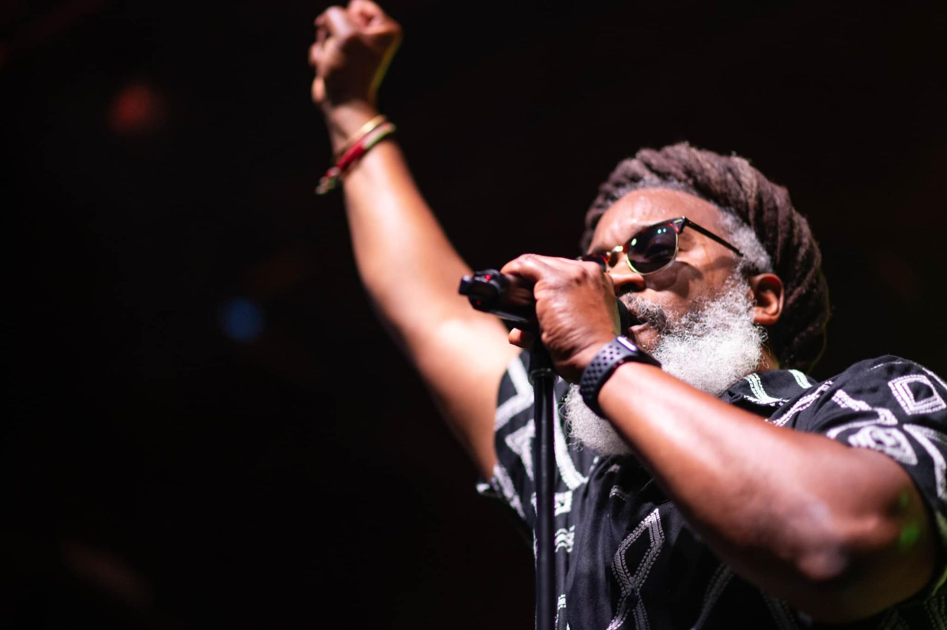 Mighty Joshua Singer with dreadlocks and sunglasses performing, arm raised, mic in hand.