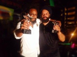 Two men holding awards, smiling. One in white shirt, glasses. Other in black. Nighttime setting.