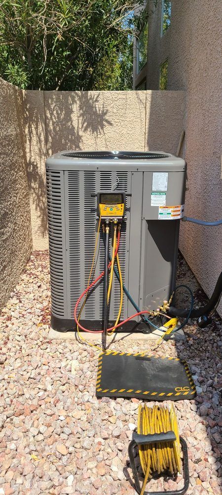 An air conditioning unit with connected gauges in a gravel-covered area. A coiled yellow extension cord sits in front.