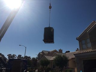 Crane lifting an air conditioning unit into the air in front of houses on a sunny day.