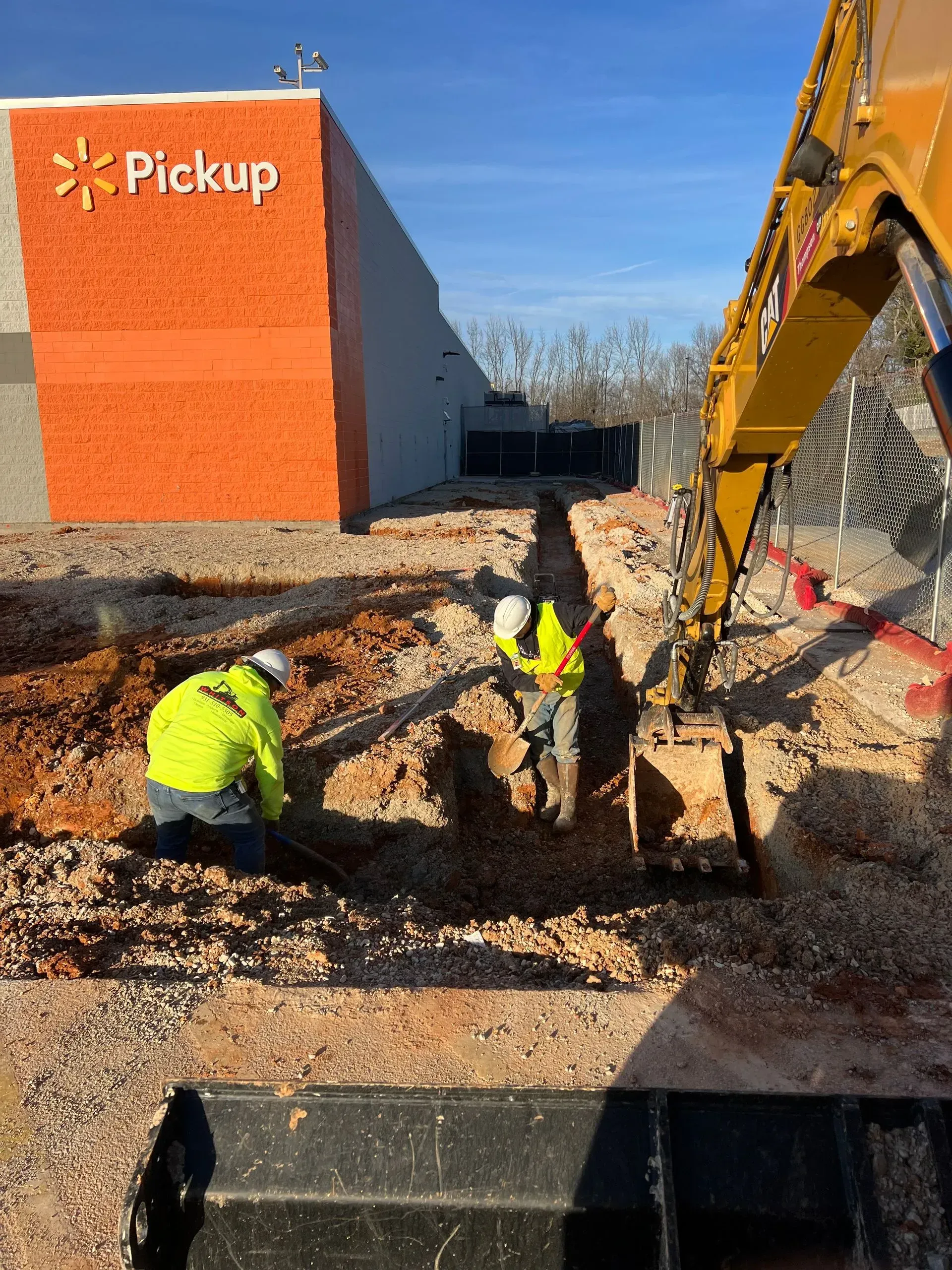 Two construction workers dig a trench near a Walmart pickup area, assisted by a large yellow excavator.