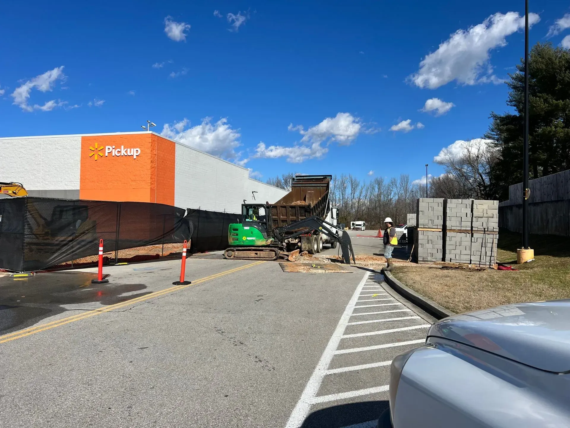 Construction workers use a small excavator to load a dump truck outside a Walmart Pickup area on a sunny day.