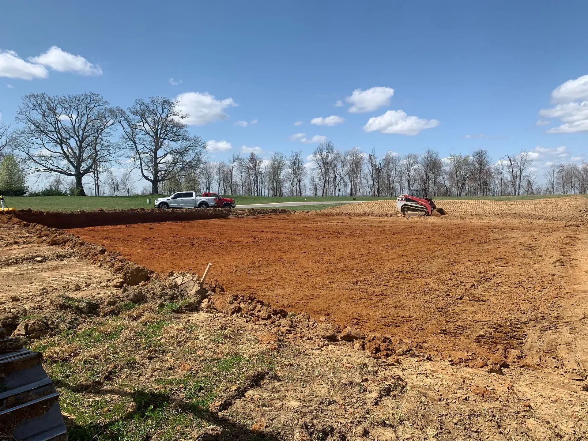 A white truck and construction equipment work on a large, excavated dirt lot against a line of trees under a blue sky.
