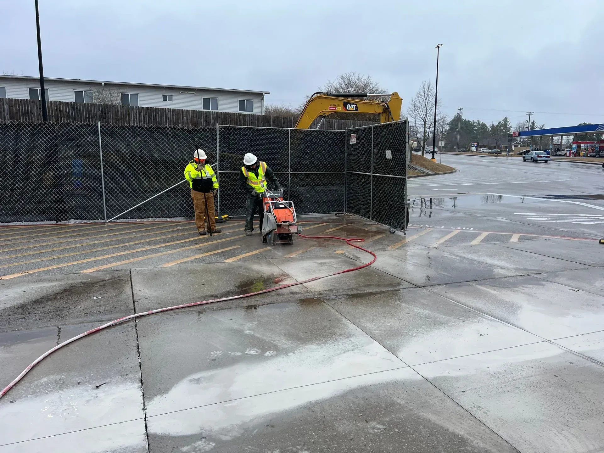 Two workers in high-visibility gear use a concrete saw on a wet parking lot near a construction fence.