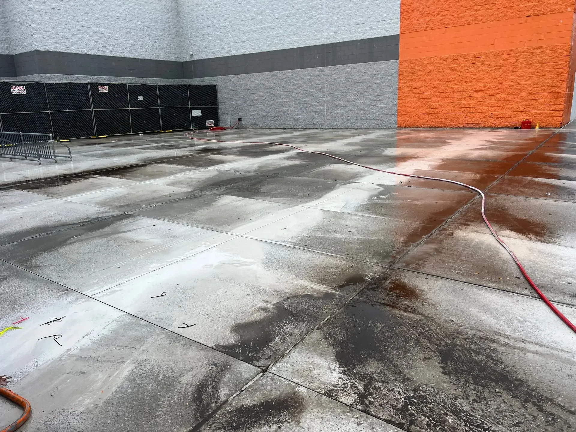 Wet concrete slab in front of a building with grey walls and an orange accent wall, with a red hose lying on the ground.