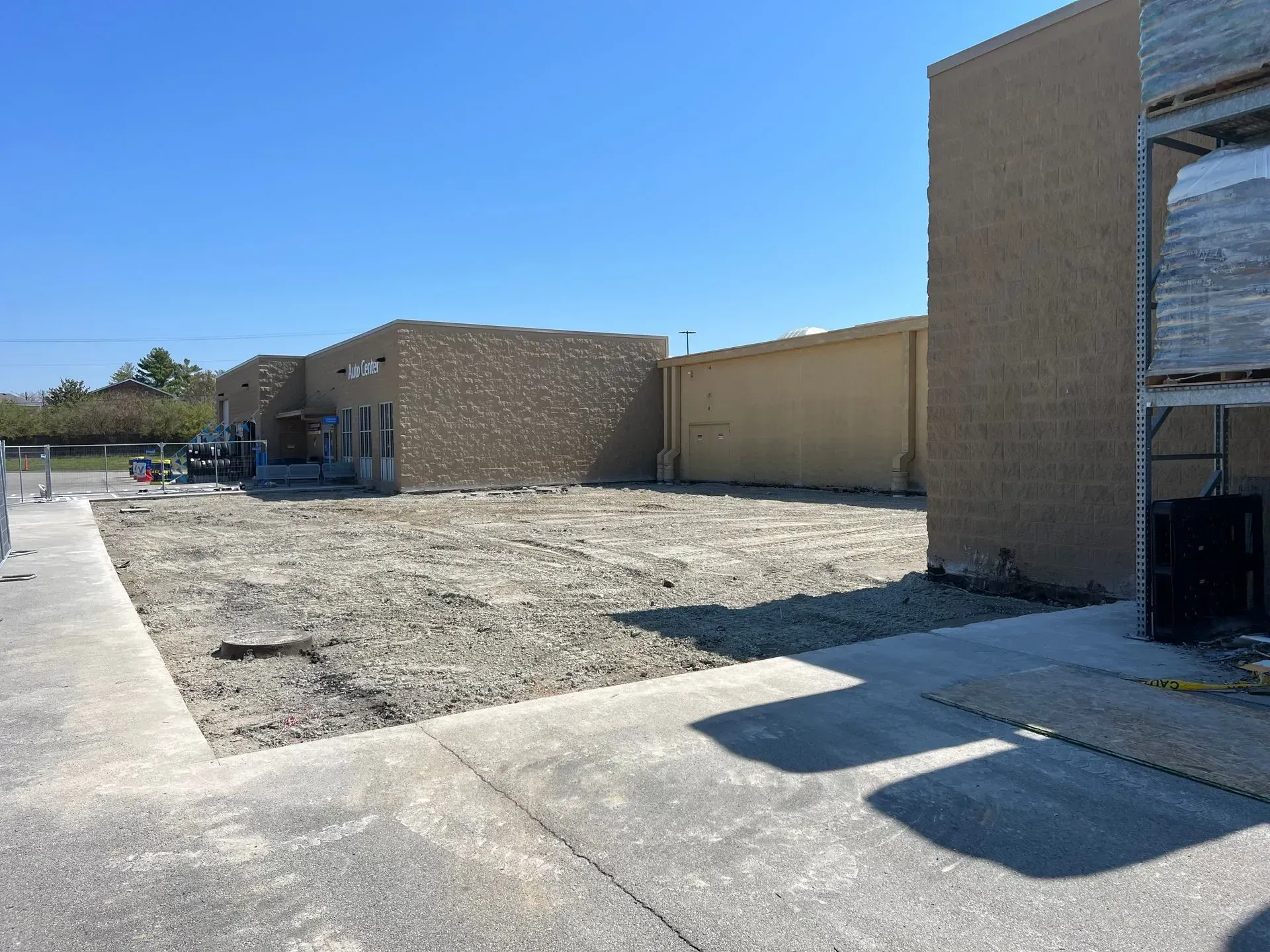 A patch of gravel and dirt sits between two tan commercial buildings under a clear blue sky on a sunny day.