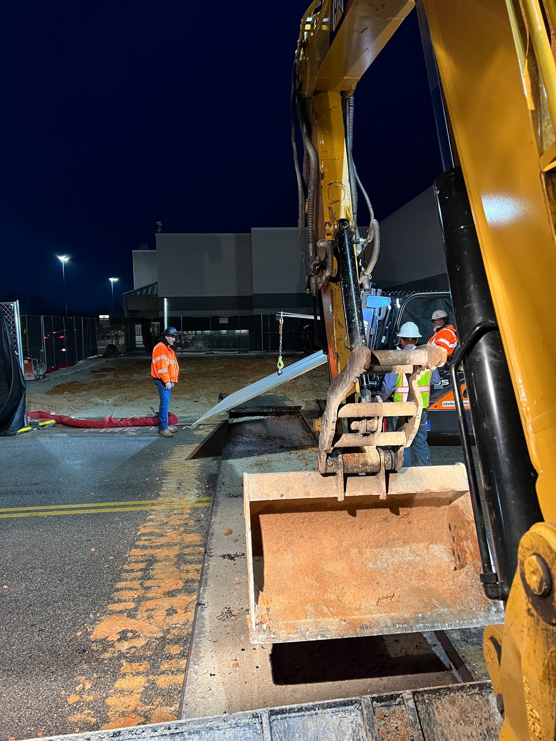 Workers in safety gear operate an excavator at night near an open trench at a construction site.