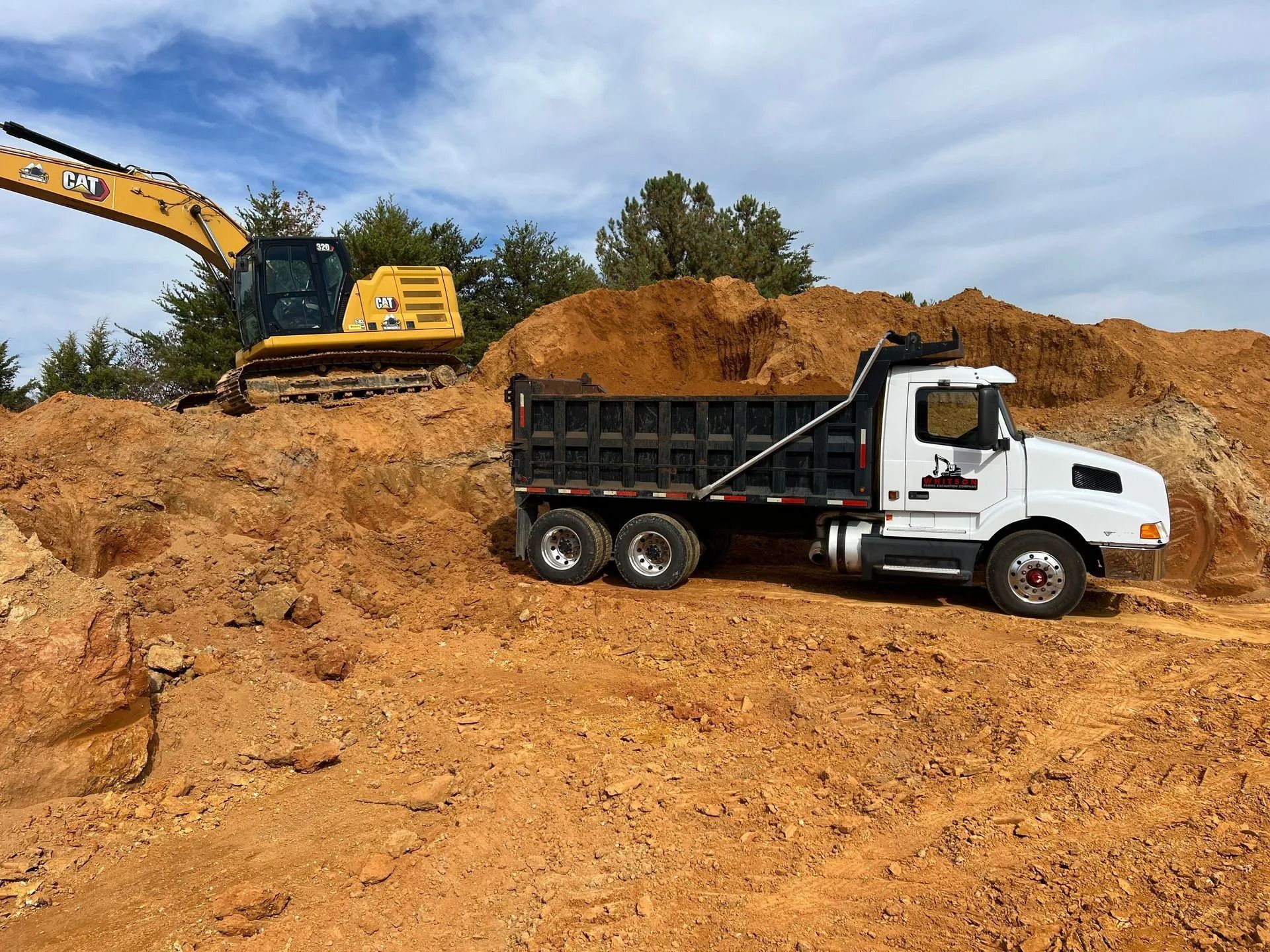 A yellow excavator sits atop a pile of dirt, positioned next to a white dump truck parked on a construction site.