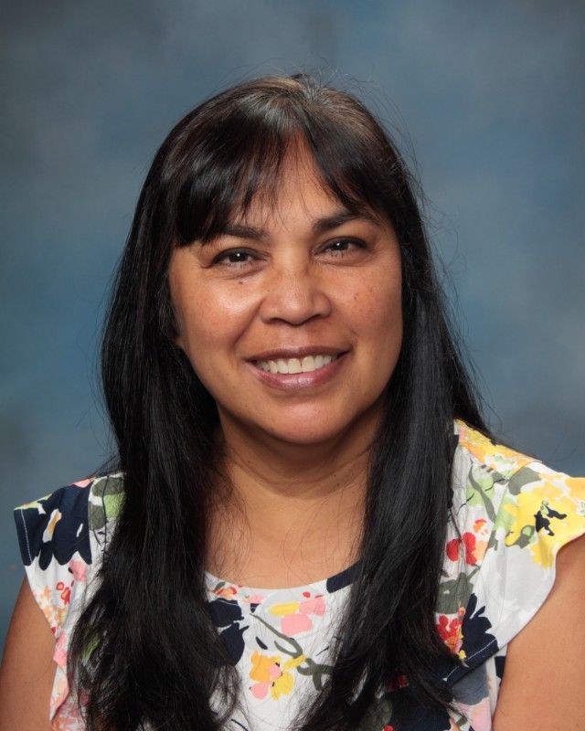 Woman with long dark hair smiles, wearing a floral top against a blue backdrop.
