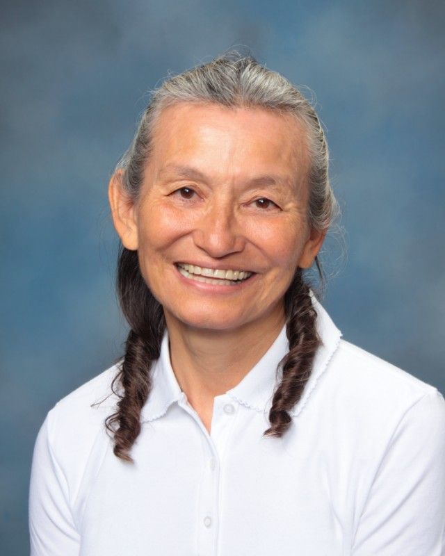 Woman with gray hair in braids smiling, wearing a white collared shirt against a blue backdrop.
