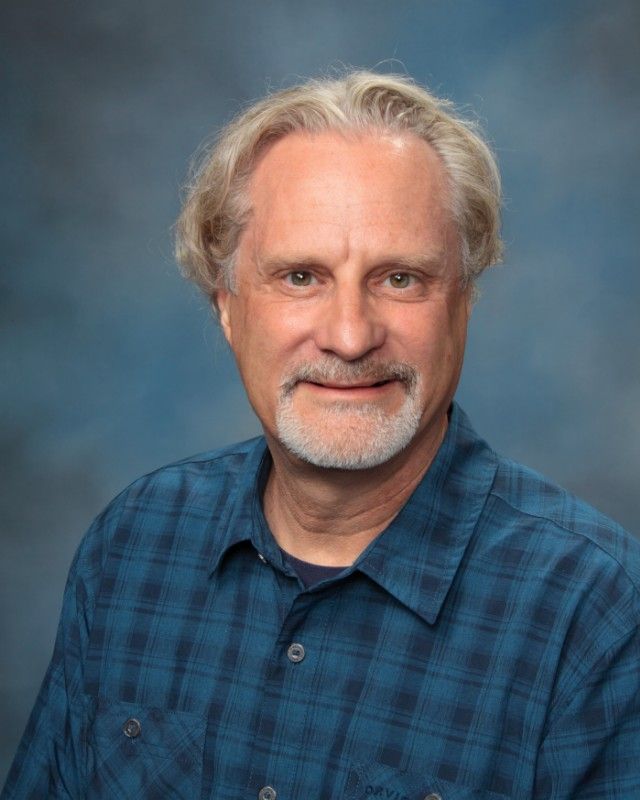 Man with graying hair and goatee, wearing a blue plaid shirt, smiling against a blue backdrop.