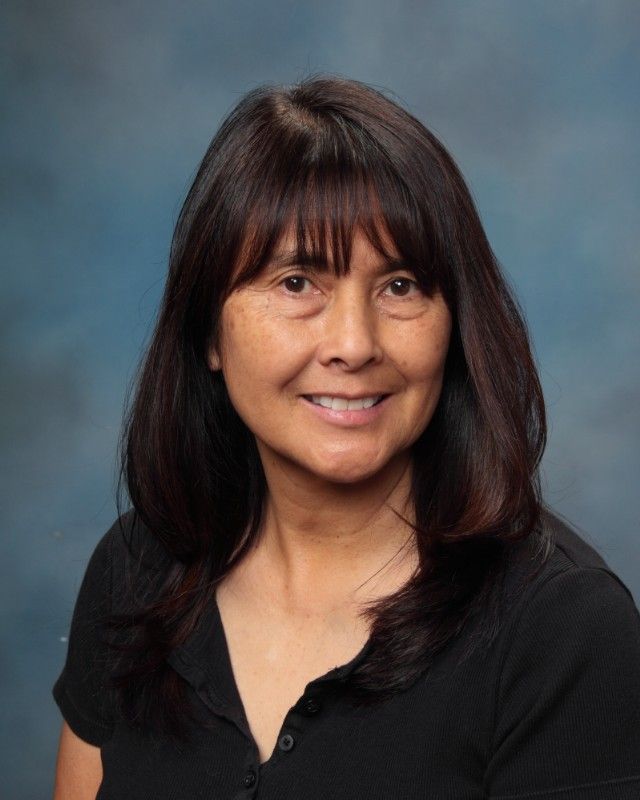 Woman with long dark hair smiles, wearing a black shirt, against a blue backdrop.