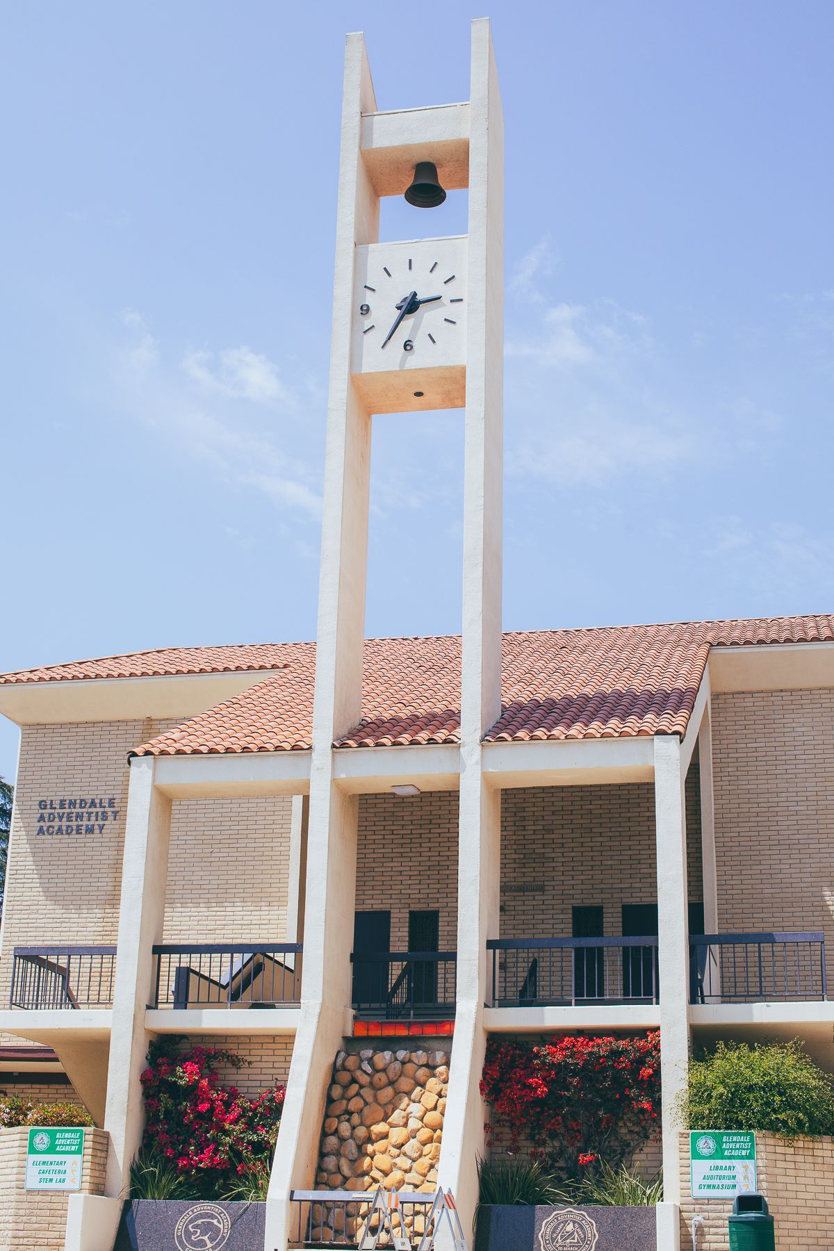 Clock tower atop a building with a tiled roof, under a blue sky; red flowers and green foliage in front.