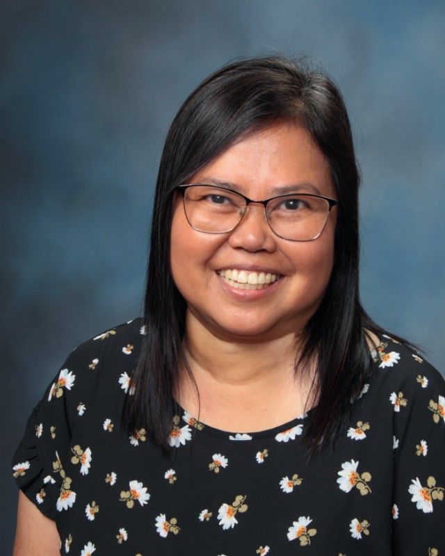 Woman with glasses smiles; dark hair, floral top, blue background.