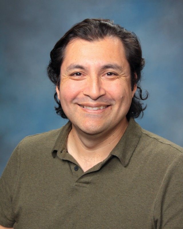 Man with dark hair, smiling, wearing an olive-green polo shirt, against a blue background.