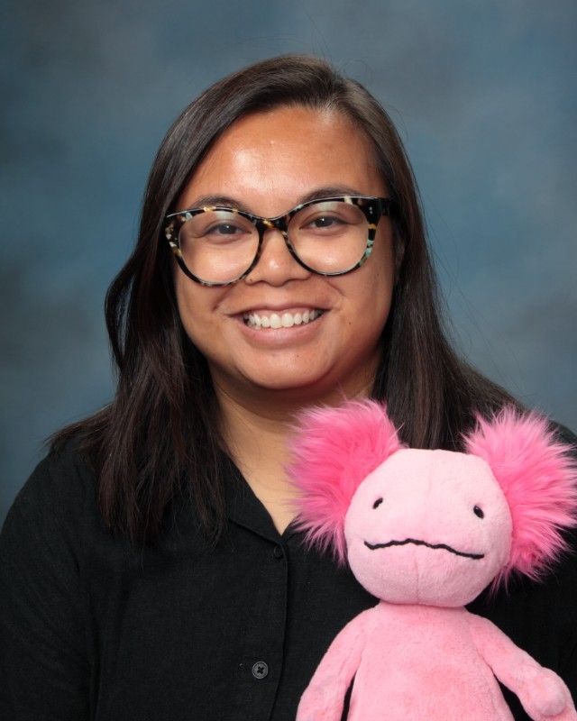 Woman with glasses smiles, holding a pink axolotl stuffed animal against a blue backdrop.