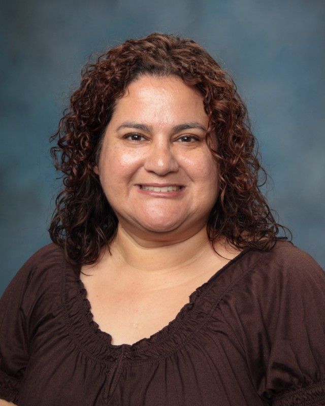 Woman with curly brown hair smiles, wearing a brown top, against a blue background.