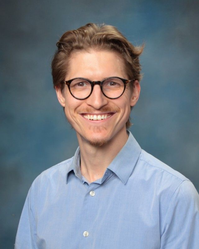 Man with glasses smiles, wearing a blue shirt, against a blurred blue background.