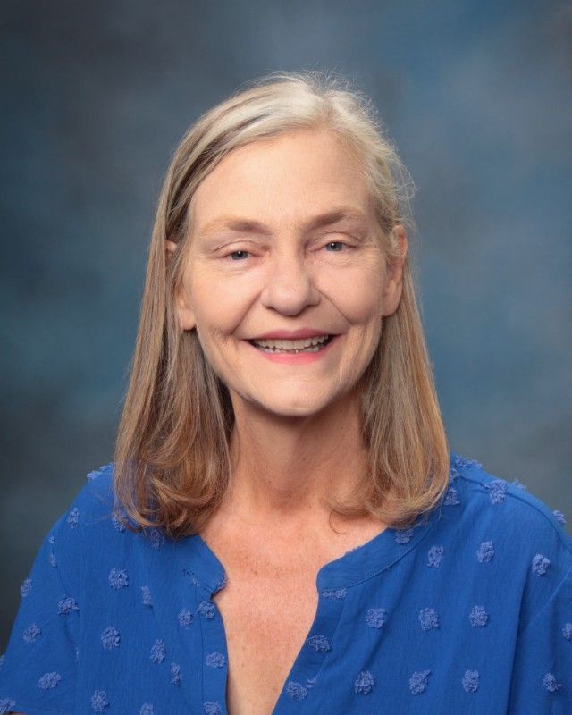 Woman with light skin smiles, wearing a blue shirt, shoulder-length blonde hair.