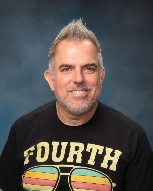 Man with grey spiky hair, wearing a black Fourth of July t-shirt, smiling, against a blue background.
