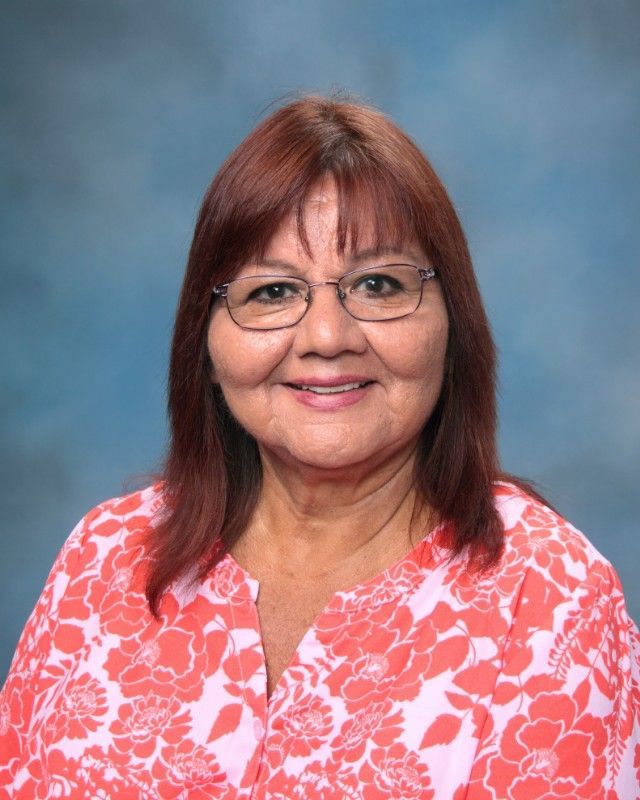 Woman with reddish-brown hair and glasses, wearing a floral orange top, smiles at the camera against a blue background.
