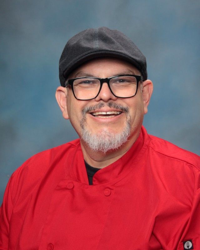 Man in a red chef's coat, glasses, and hat smiles broadly, in front of a blue background.