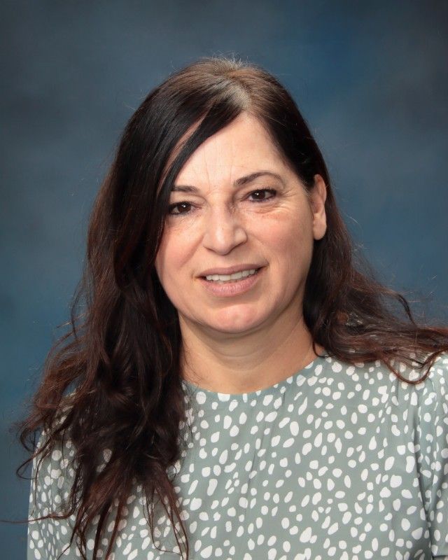 Woman with long dark hair smiles, wearing a green and white patterned top against a blue background.
