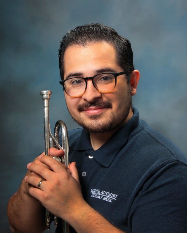 Man holding a trumpet, wearing glasses and a dark blue shirt with logo; smiling against a blue backdrop.