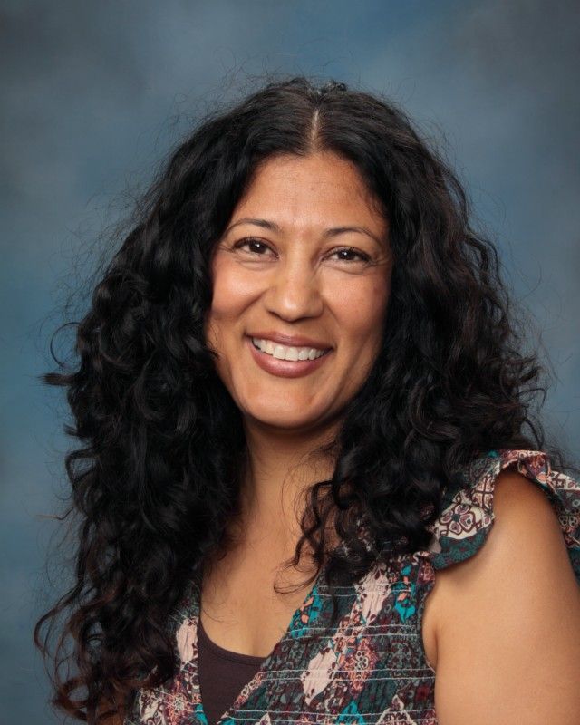Woman with dark curly hair smiles at the camera, wearing a floral top against a blue background.