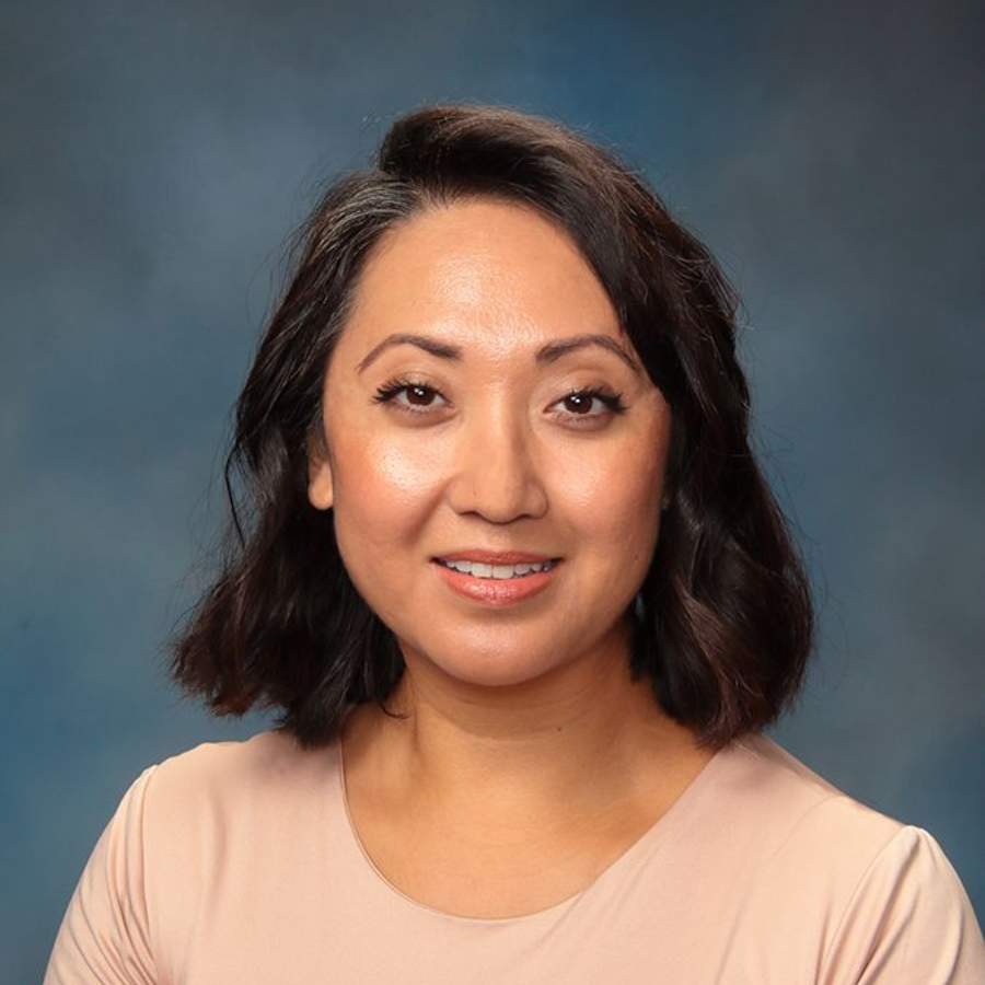 Woman with dark hair smiles, wearing a pink top against a blue backdrop.
