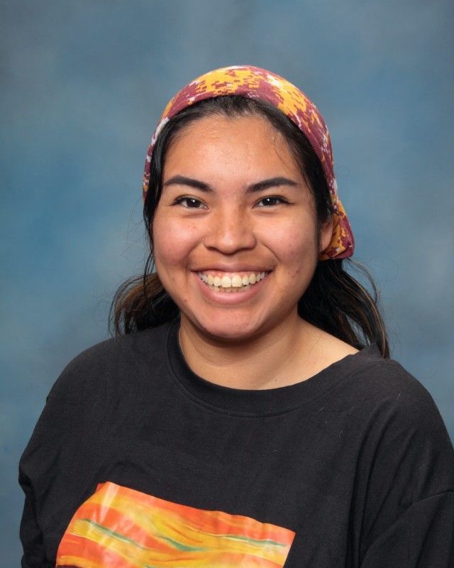 Woman with a bright smile wearing a black shirt and a colorful headscarf, against a blue background.