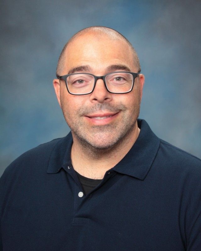 Bald man wearing glasses and a navy blue polo shirt, smiling slightly against a blue backdrop.