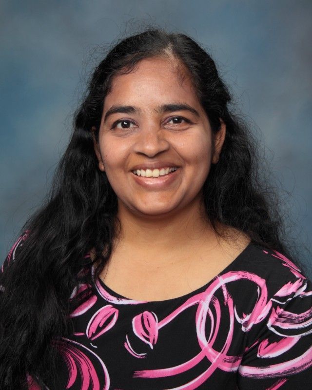 Woman with long dark hair, smiling, wearing a black and pink patterned top, against a blue background.
